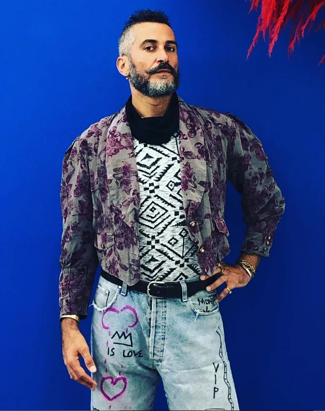 Marco Scola wearing hand-painted upcycled jeans and jacket against a blue wall.